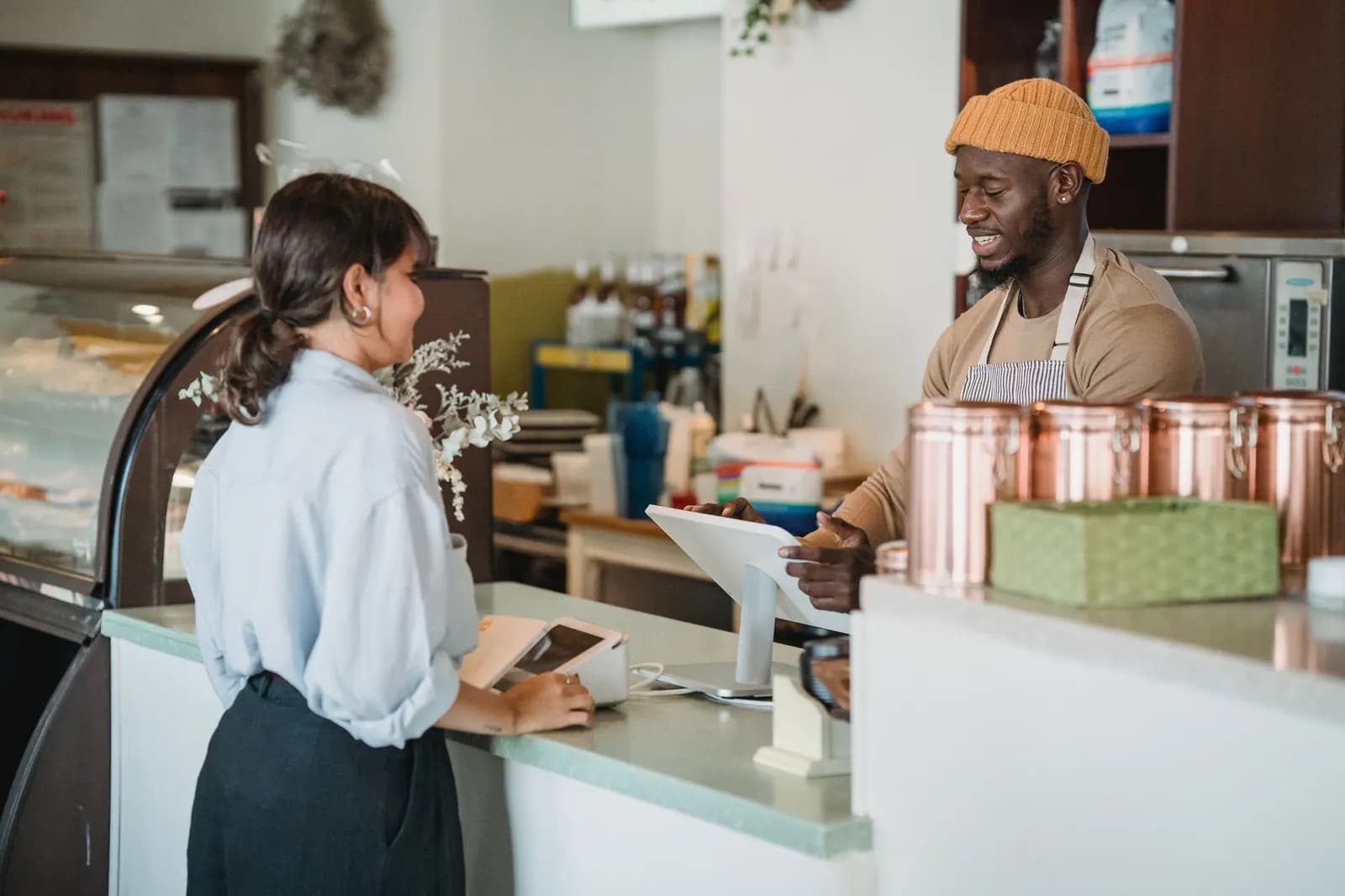 Barista και πελάτισσα συζητούν χαμογελαστοί στο ταμείο μιας φωτεινής καφετέριας