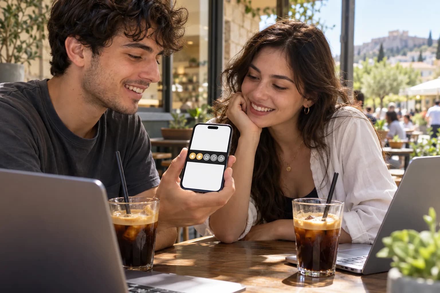 Δύο φοιτητές σε καφέ με freddo espresso και laptop κοιτούν μια κάρτα σφραγίδων στο κινητό